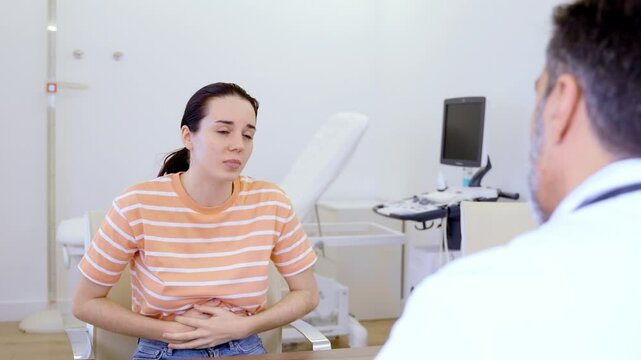 Concerned female patient describing abdominal pain symptoms during medical consultation with attentive physician in clinical examination room