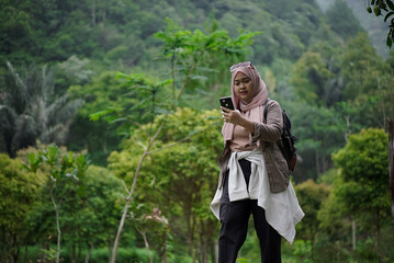 Young Indonesian woman wearing hijab walking in nature while taking photos of natural scenery, enjoying the scenery and natural atmosphere, travel concept.
