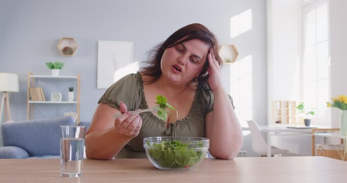 Sad fat plus-size woman eats a vegetable salad at home living room. Her diet focuses on healthy food, though her dissatisfaction is evident as she engages in this health-conscious routine.