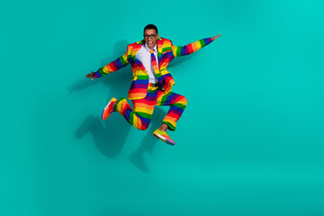 Charismatic man in vibrant rainbow-striped suit enthusiastically jumping against bright turquoise...