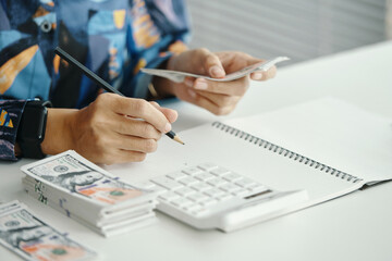 Hands of unrecognizable female accountant holding pencil and bundle of money, writing in notebook