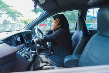 A woman in a black hijab is driving a car and covering his ears with his hands. She is wearing a black head scarf and she is in a hurry