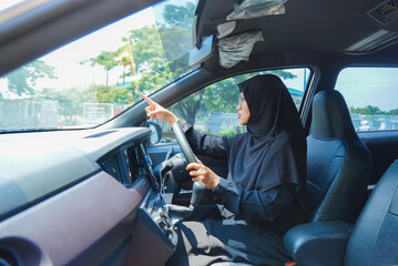 A woman in a black hijab is driving a car and pointing to the right. The car is parked in a parking lot