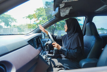 A woman in a black hijab is driving a car and pointing to the right. The car is parked in a parking lot