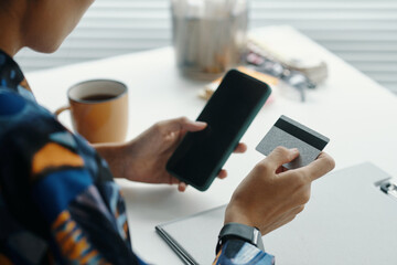 Over shoulder shot of unrecognizable young woman doing online shopping using app on smartphone and bank card, while having break at work