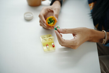 From above view of unrecognizable female hands adding vitamin capsules into pill organizer, while sitting at desk