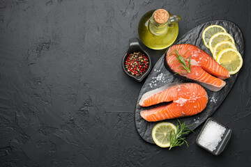 Fresh raw salmon steaks with salt, lemon, oil, peppercorns and rosemary on dark textured table, flat lay. Space for text