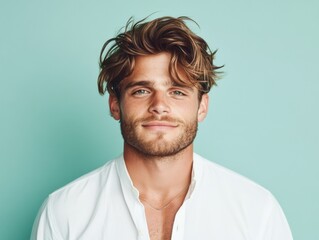 Obraz premium A smiling young man with tousled hair in a white shirt stands against a light blue background, exuding a relaxed and confident aura.