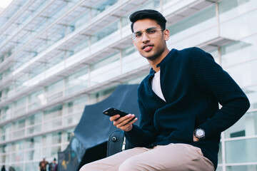 Portrait of handsome hindu male in stylish wear and spectacles using mobile phone for chatting and...