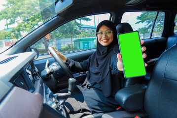 A woman in a black hijab is holding a green phone in a car. She is smiling and she is happy