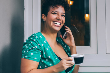 Joyful woman speaking on smartphone with cup in hand at table in outside cafe