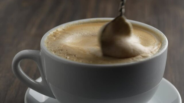 Stirring foam of hot coffee cappuccino with spoon in white cup on wooden table, close up. Milk foam cappuccino, latte