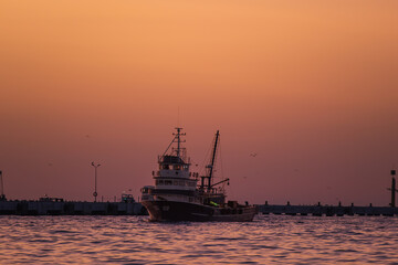 Fototapeta premium A fishing boat docked at a harbor during a stunning orange sunset, with seagulls flying over the tranquil sea.