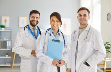 Group of happy doctors standing together in medical office, looking directly at camera. Friendly...