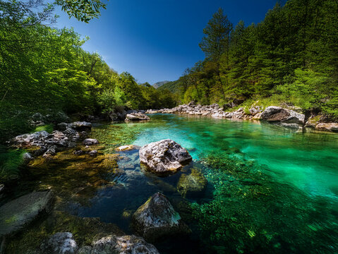 A serene river flows through a lush green valley under a vibrant blue sky.  Sunlight illuminates the crystal-clear water, revealing the riverbed below.  Rocks line the banks, adding to the natural bea