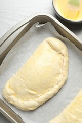 Uncooked calzone pizza in baking dish and egg yolk on white wooden table, top view