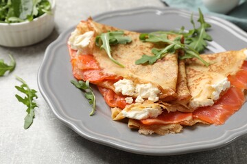 Delicious crepes with salmon, cream cheese and arugula on grey table, closeup