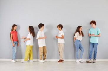 Diverse happy smiling children in casual wear leaning on grey wall studio background and talking. Full body length indoor group portrait of preteen boys and girls standing together, making friends.