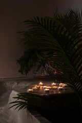 A tranquil arrangement of tea light candles on a concrete ledge surrounded by lush green palm leaves. The warm glow of the candles contrasts beautifully with the dark, moody background, evoking a sens