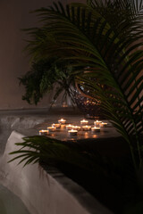 A tranquil arrangement of tea light candles on a concrete ledge surrounded by lush green palm leaves. The warm glow of the candles contrasts beautifully with the dark, moody background, evoking a sens © Юлия Безуглая