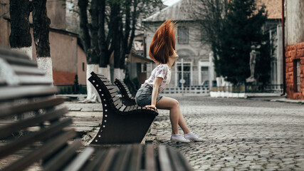 The girl leaned back, her long red hair flying in the air. The bench she sits on has a modern design with metallic elements.