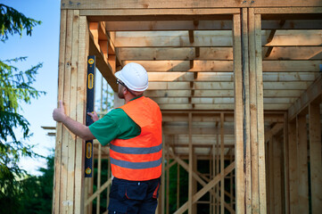 Carpenter constructing two-storey wooden frame house. Man inspects walls for levelness using spirit level, wearing protective overalls, helmet and orange vest. Concept of contemporary construction.