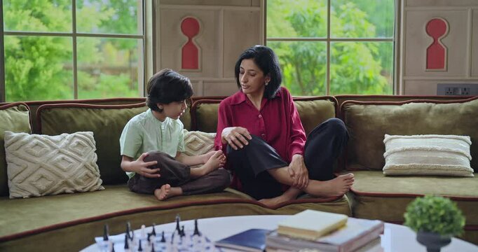Indian happy relax young adult female lady with cute little son boy kid child teach yoga asana home family sitting on cozy sofa couch talk learn good body health care habit have joy fun enjoy day time