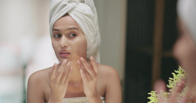 Indian upset young adult gen z woman lady wrapped towel on head and body look mirror feel sad facial pores skin dry cheek teen girl female worried for acne pimple touch face heath care at indoor home