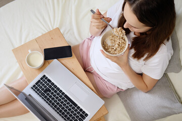 A woman sits comfortably on a bed, eating a bowl of oatmeal. She balances her laptop on her lap and drinks from a cup, starting her day with a productive morning routine
