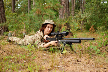A female soldier lies prone on the ground, focusing intently on a long-range sniper rifle in a...
