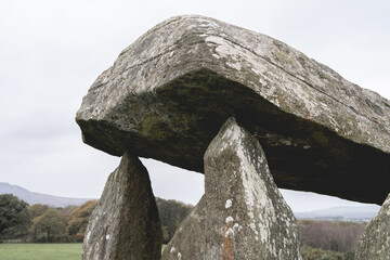 A weathered dolmen with a flat stone atop three upright stones sits in a lush green landscape,...