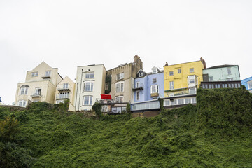 Picturesque row of colorful houses on a steep hillside, featuring bay windows, balconies, and varying rooflines, with vibrant hues contrasting against lush greenery and a cloudy sky.