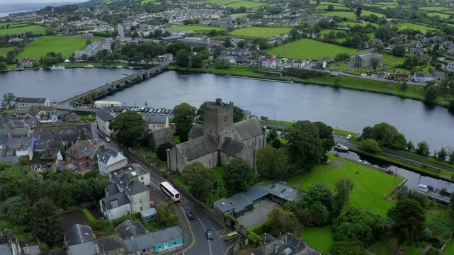 St Flannan's Cathedral, Killaloe, County Clare, Ireland, September 2021. Drone orbits counter clockwise from a high angle with Shannon River bordering the old cemetary as cars drive through the town.
