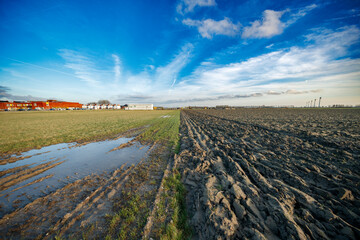 Plowed field and grassy area.  Buildings in background, clear blue sky.  Muddy tracks and puddles.