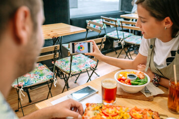 Young girl showing fresh poke bowl meal photo on smartphone to friend eating vegetarian pizza and...