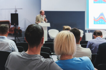 Group of people are sitting in a room and watching a presentation. The man giving the presentation is standing in front of a podium. The audience appears to be attentive and engaged