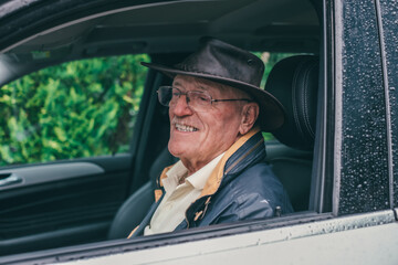 Portrait of smiling senior man with leather hat sitting on driver seat in his white car while it's raining. Old caucasian man on driving vehicle in a rainy day for transport and copy space