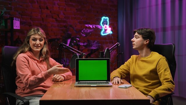 Man host and woman guest recording interview in loft podcast studio using microphones, talking creating digital content, laptop with chroma key green screen mockup.