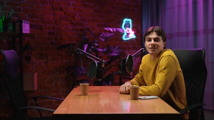 Man host sitting at the table looking talking at camera, recording interview in loft podcast studio...