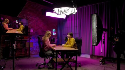 Backstage of man host and woman guest recording interview in loft podcast studio using microphones, creating digital content, presenter and woman sit at table and start conversation.