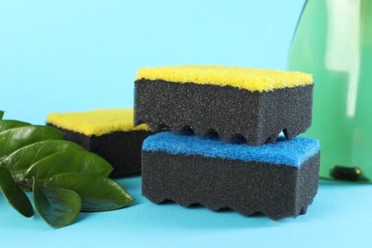 Many colorful sponges and branch with leaves on light blue background, closeup - Powered by Adobe