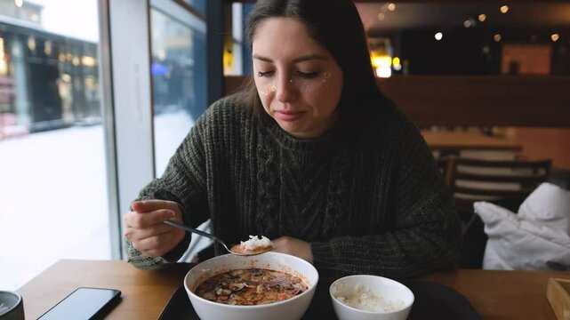 Pretty brunette enjoying delicious Tom Yum Kun soup with shrimps in Thai restaurant winter in Siberia. A woman in a warm cozy sweater is having lunch near the window. Snow outside