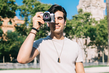 Happy caucasian male traveler holding camera focusing while taking picture of urban setting in city, cheerful man photographer amateur enjoying spending free time on hobby during trip vacations