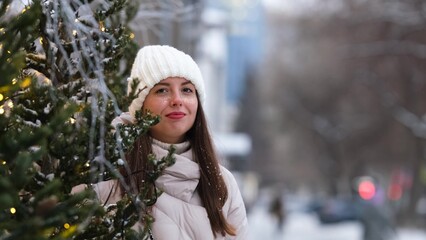 Portrait of a beautiful young woman in winter clothes. The woman smiles happily against the background of a snowy street traditionally decorated for Christmas holidays