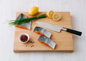 Fresh salmon fillets prepared with lemon and herbs on a wooden cutting board complemented by lemon halves, chives, and a bowl of colorful peppercorns, ready for cooking a nutritious meal