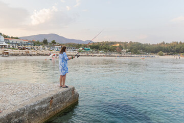 Woman fishing on a pier, gazing over a serene beach resort, calm waters, and distant hills.