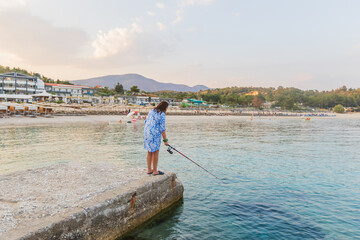 Naklejka premium Woman fishing on a pier, gazing over a serene beach resort, calm waters, and distant hills.