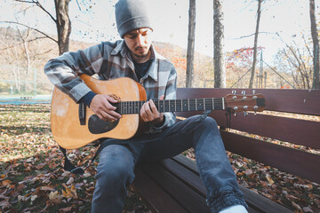 Naklejka premium Man playing acoustic guitar outdoors in autumn, wearing a plaid shirt, sitting on a wooden bench surrounded by fallen leaves. Perfect for themes of music, relaxation, and nature
