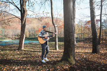 Man wearing a plaid shirt and a beanie stands in a serene autumn park, strumming an acoustic guitar. Surrounded by colorful leaves and tall trees, he expresses creativity and passion for music outdoor