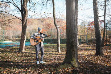 Man wearing a plaid shirt and a beanie stands in a serene autumn park, strumming an acoustic guitar. Surrounded by colorful leaves and tall trees, he expresses creativity and passion for music outdoor